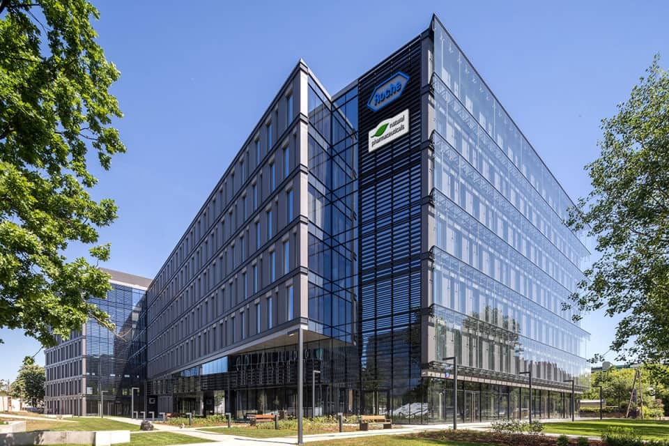 Natural Pharmaceuticals Movisan™ Collagen II Modern üvegfelületű irodaház zöld fák között, két vállalati logóval a homlokzaton kék égen.