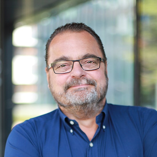 Natural Pharmaceuticals Movisan™ Collagen II Középkorú férfi szemüvegben, kék ingben, mosolyogva, beltéri, elmosódott üvegfalas háttér előtt.