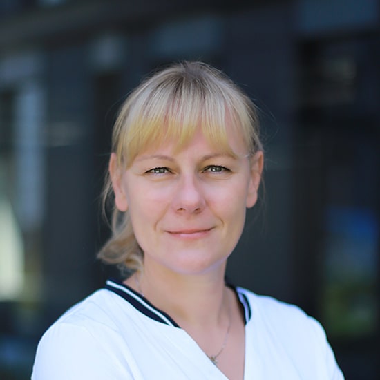 Natural Pharmaceuticals Movisan™ Collagen II Közeli kép egy mosolygó, világos bőrű, szőke nő portréjáról, elmosódott városi háttérrel.