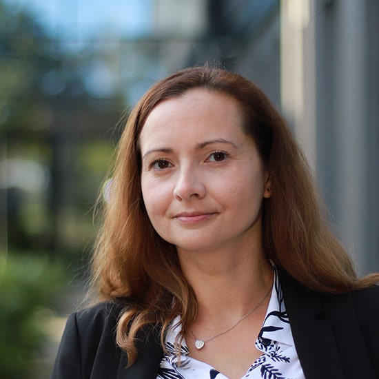 Natural Pharmaceuticals Movisan™ Collagen II Barna hajú nős nő fekete zakóban, enyhén mosolyogva, elmosódott városi háttér előtt.