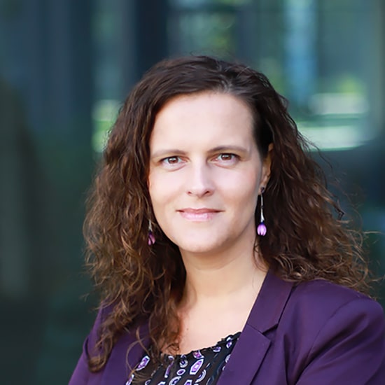 Natural Pharmaceuticals Movisan™ Collagen II Barna hajú nő lila zakóban, hullámos hajjal és lila fülbevalóval, homályos háttér előtt áll.