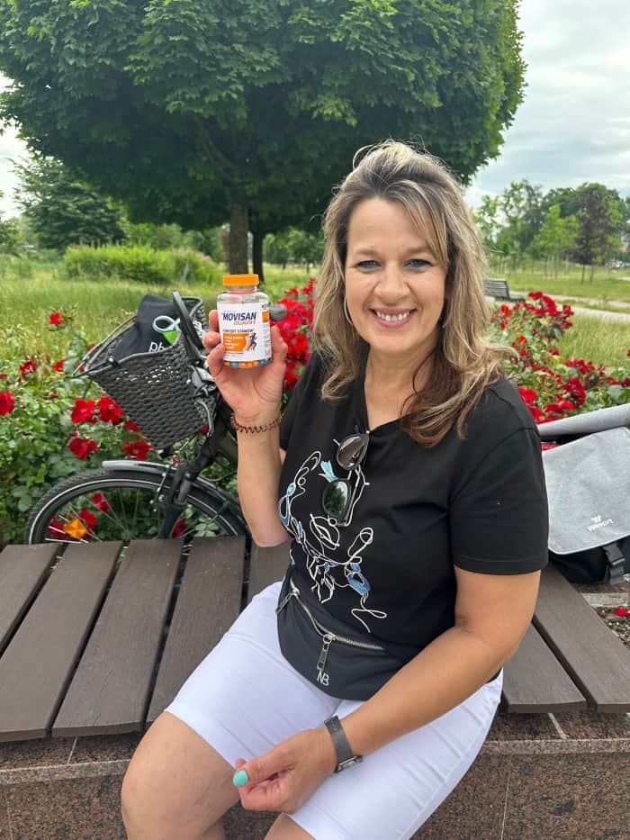 Natural Pharmaceuticals Movisan™ Collagen II Mosolygó nő ül a parkban, kezében mozgásszervi étrend-kiegészítőt tart, kerékpár és virágok háttérben.