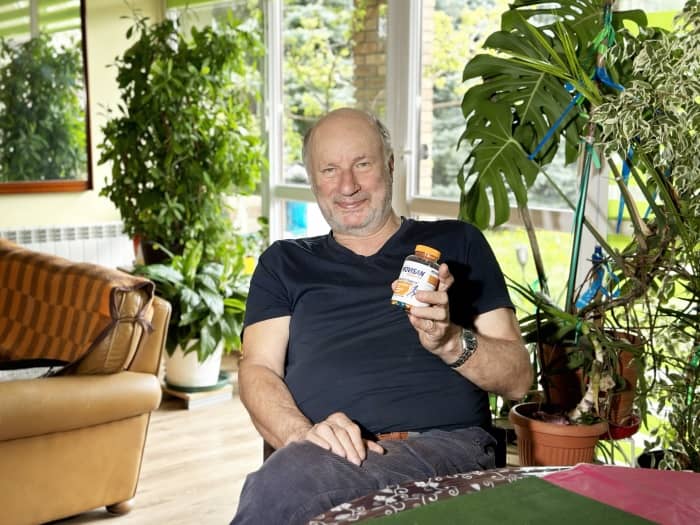 Natural Pharmaceuticals Movisan™ Collagen II Mosolygó férfi otthoni növények között, kezében étrend-kiegészítő tablettás üveggel.