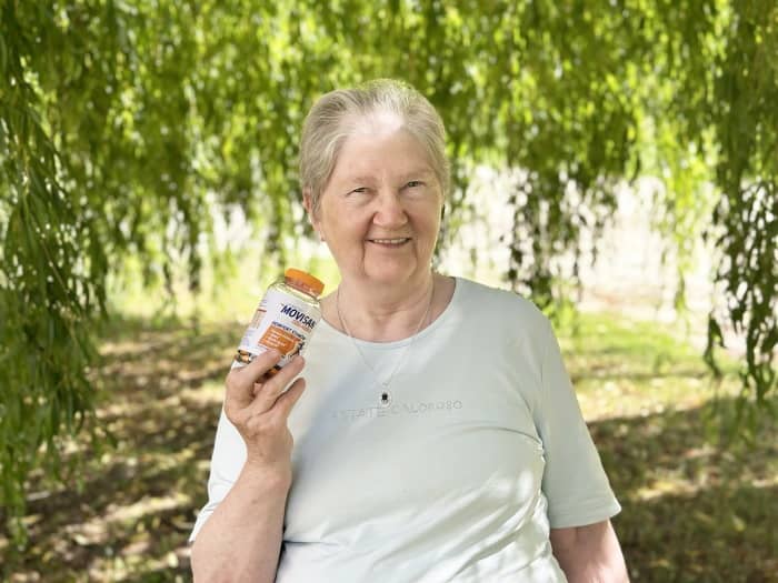Natural Pharmaceuticals Movisan™ Collagen II Idős nő mosolyogva tartja kezében a Movisca ízületvédő kapszulákat zöld parkban.
