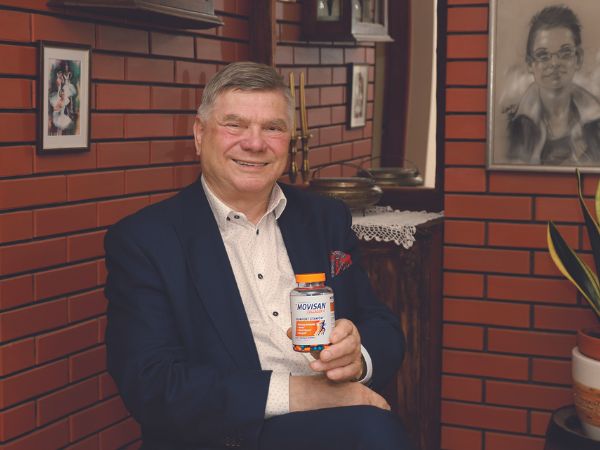 Natural Pharmaceuticals Movisan™ Collagen II Mosolygó idős férfi Movisan ízületvédő kapszulával téglafal előtt ülve, otthoni környezetben.