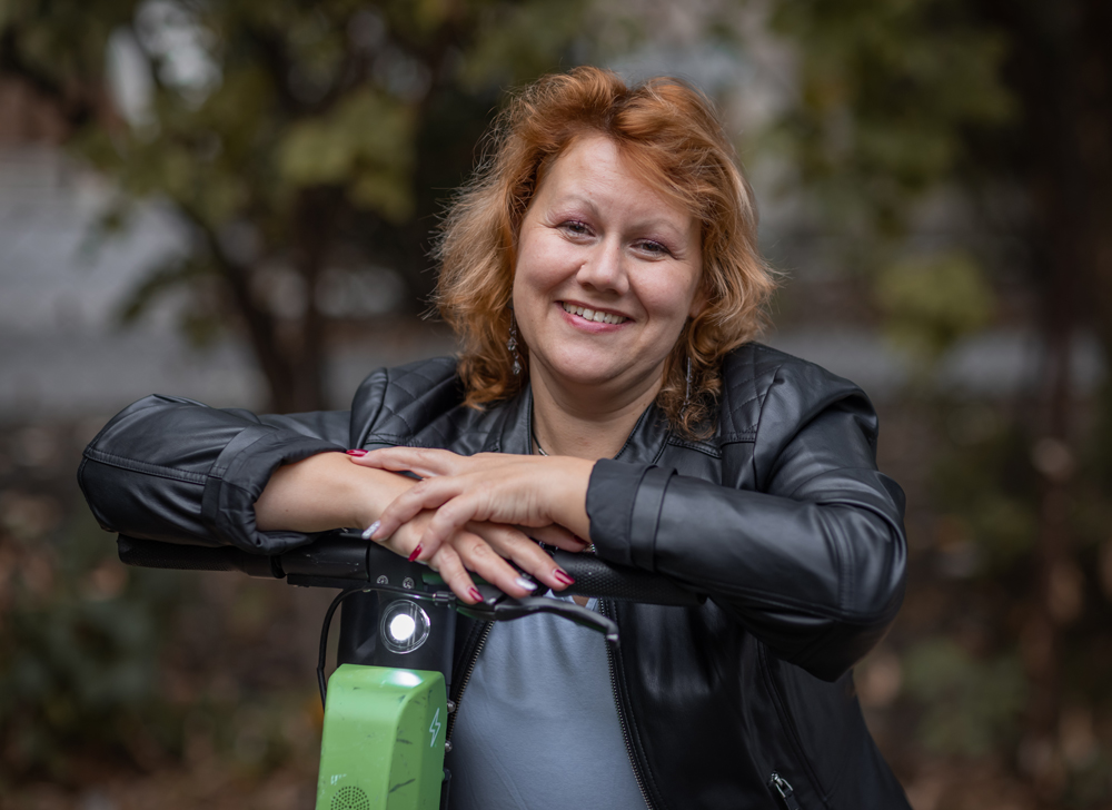 Natural Pharmaceuticals Movisan™ Collagen II Mosolygó nő fekete bőrdzsekiben elektromos rollerrel parkban, őszi háttérrel.